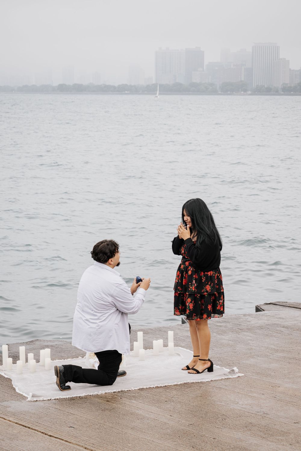 Man proposes on one knee to woman on waterfront dock with candles and city skyline backdrop.