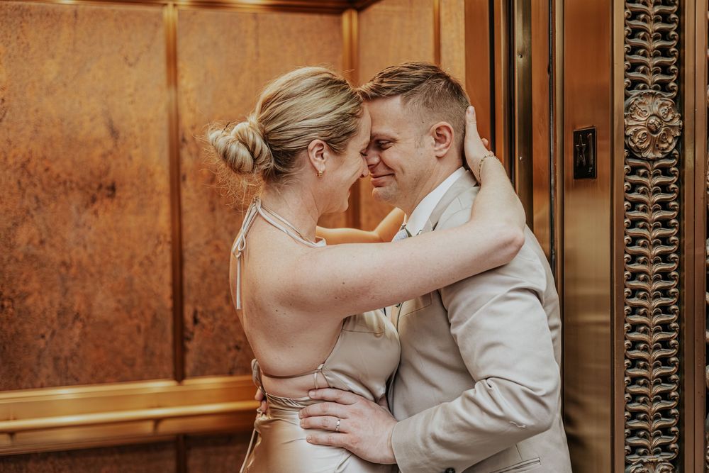 A couple in wedding attire embraces and smiles at each other in front of an ornate golden elevator.