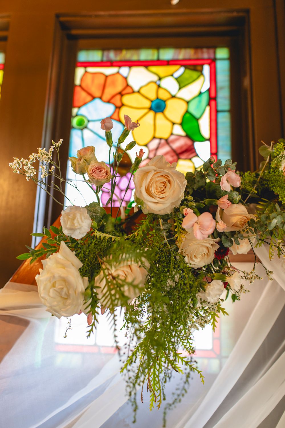 Elegant floral arrangement with roses and greenery against a colorful stained glass window.