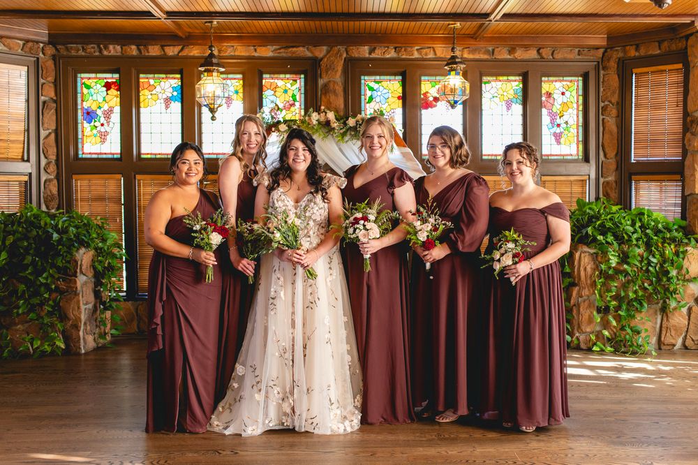 A bridal party poses in burgundy dresses against a wooden wall with stained glass windows.