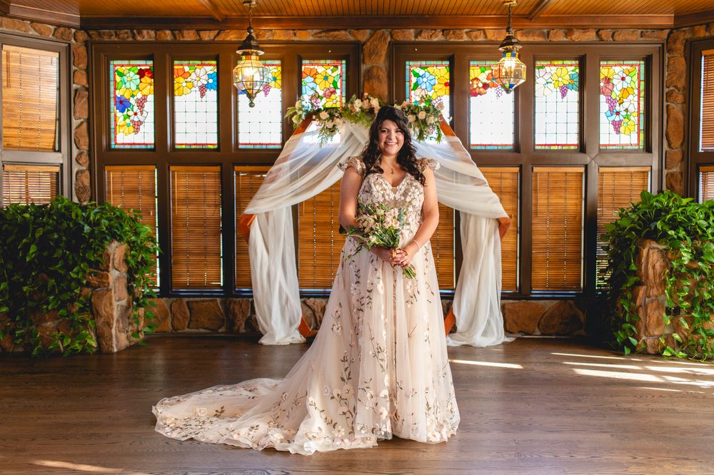 A wedding dress with lace detailing is showcased in front of stained glass windows and white drapes.