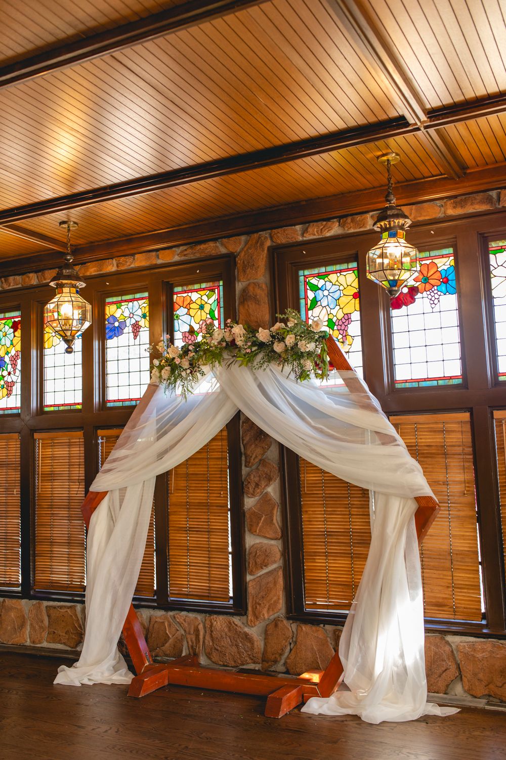 White wedding arch with floral arrangements against wooden walls with stained glass windows.