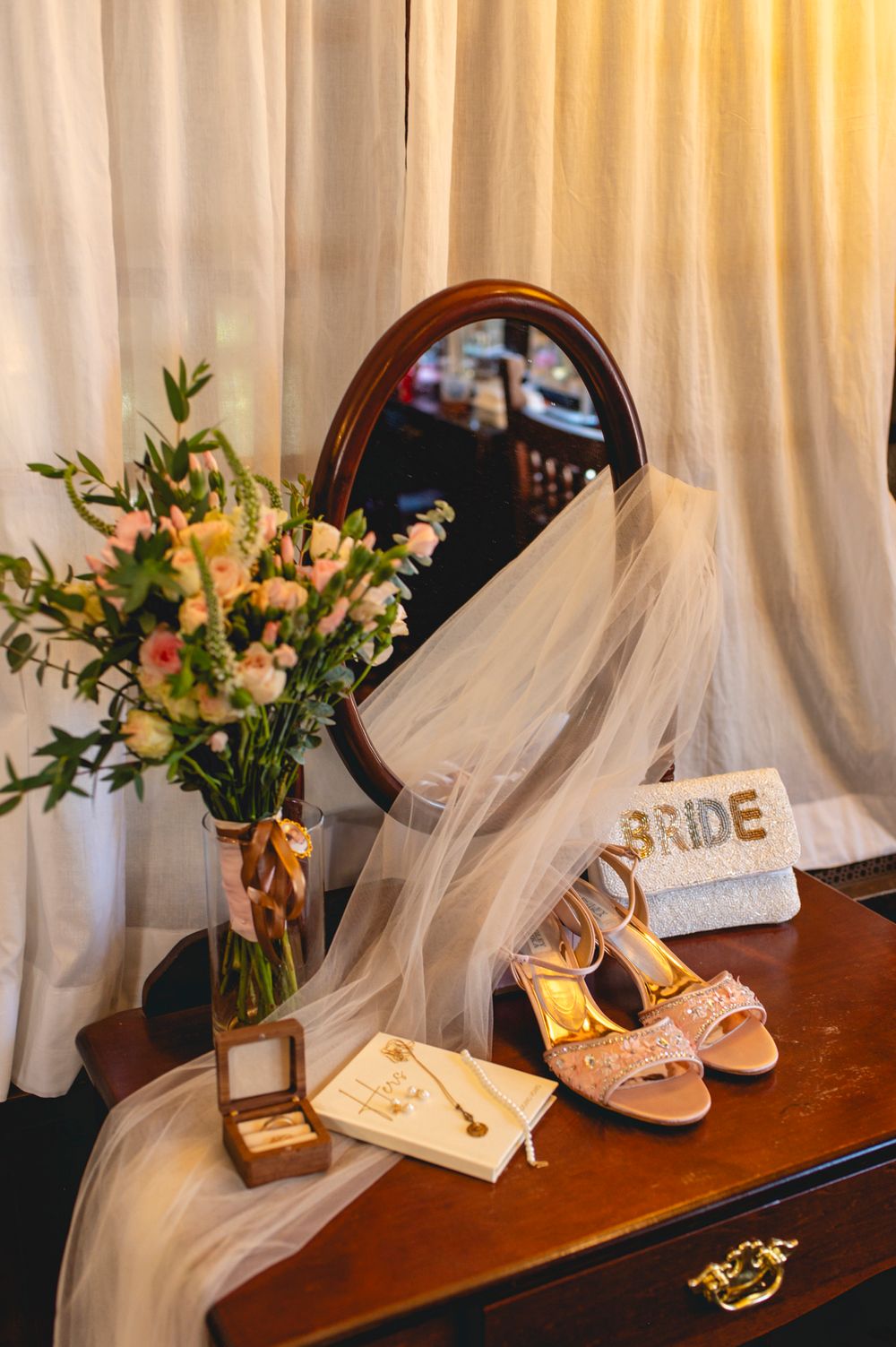 Wedding accessories including bridal shoes, veil and floral arrangement displayed on a wooden dresser with mirror.