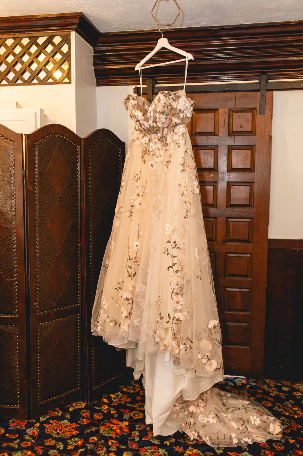 An elegant champagne-colored wedding dress with floral appliques hanging on display in a wood-paneled room.