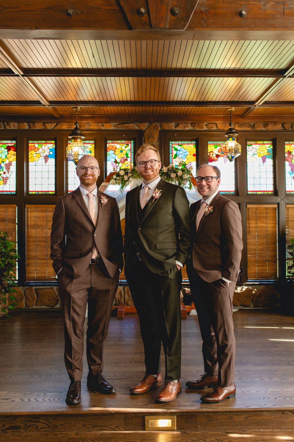 Three groomsmen in suits pose in front of stained glass windows in a rustic venue.
