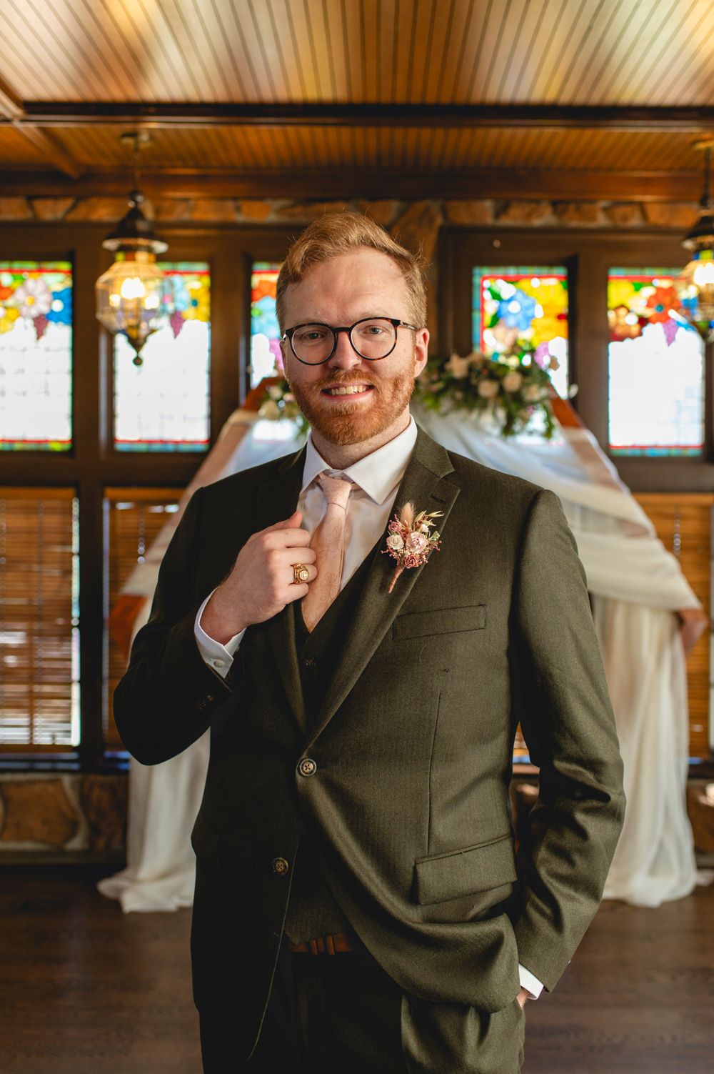 A groom in olive green suit and glasses poses in front of stained glass windows.
