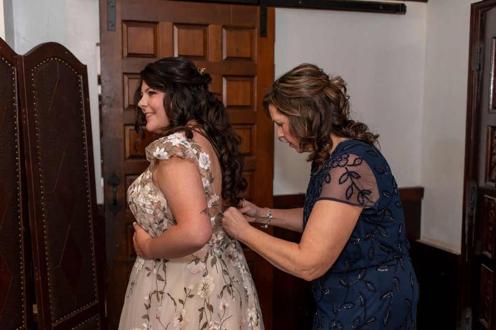 Two people in formal dresses during wedding preparations in a wood-paneled room.