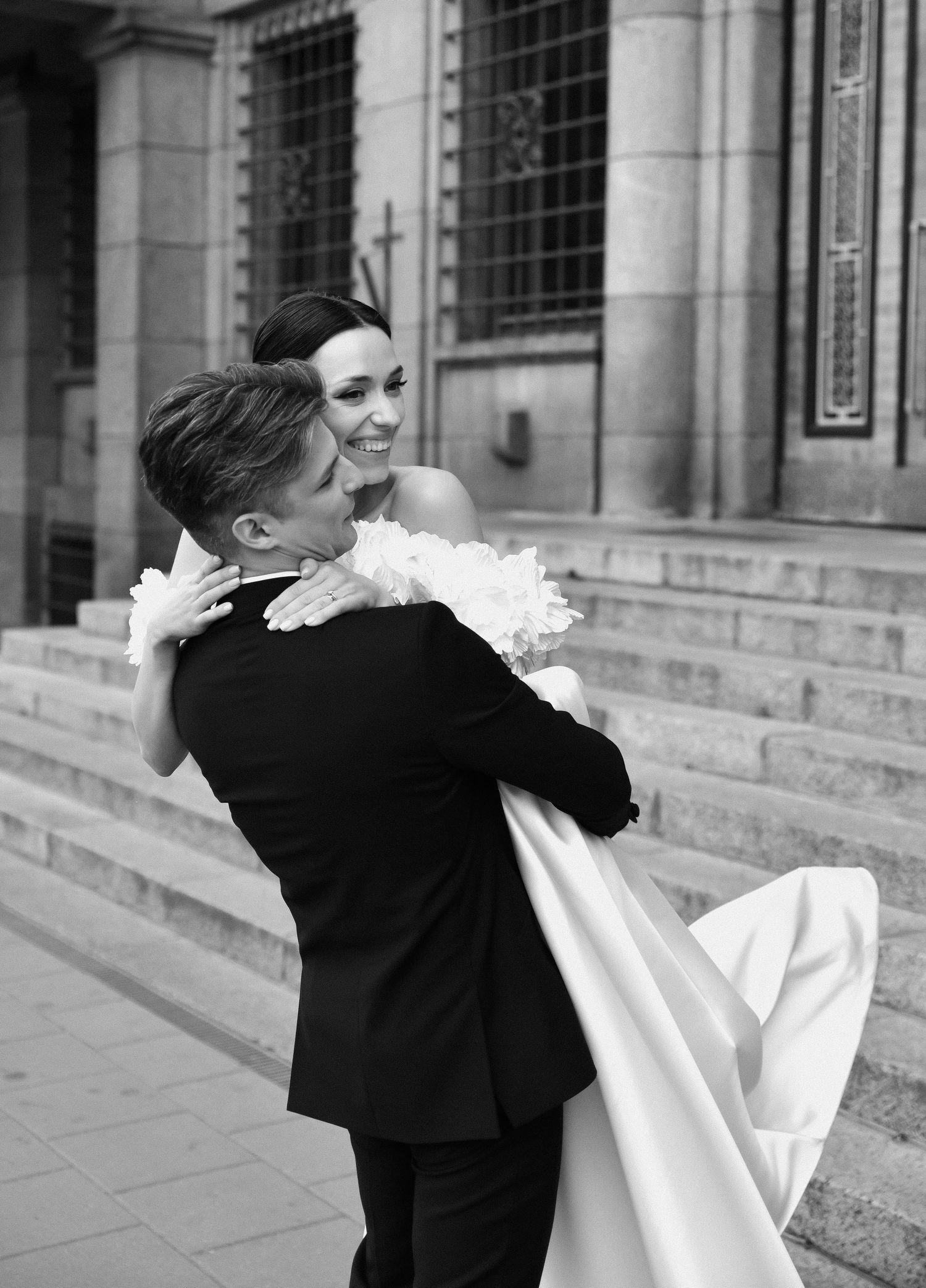 Wedding couple poses against lush greenery backdrop holding bouquet and champagne.