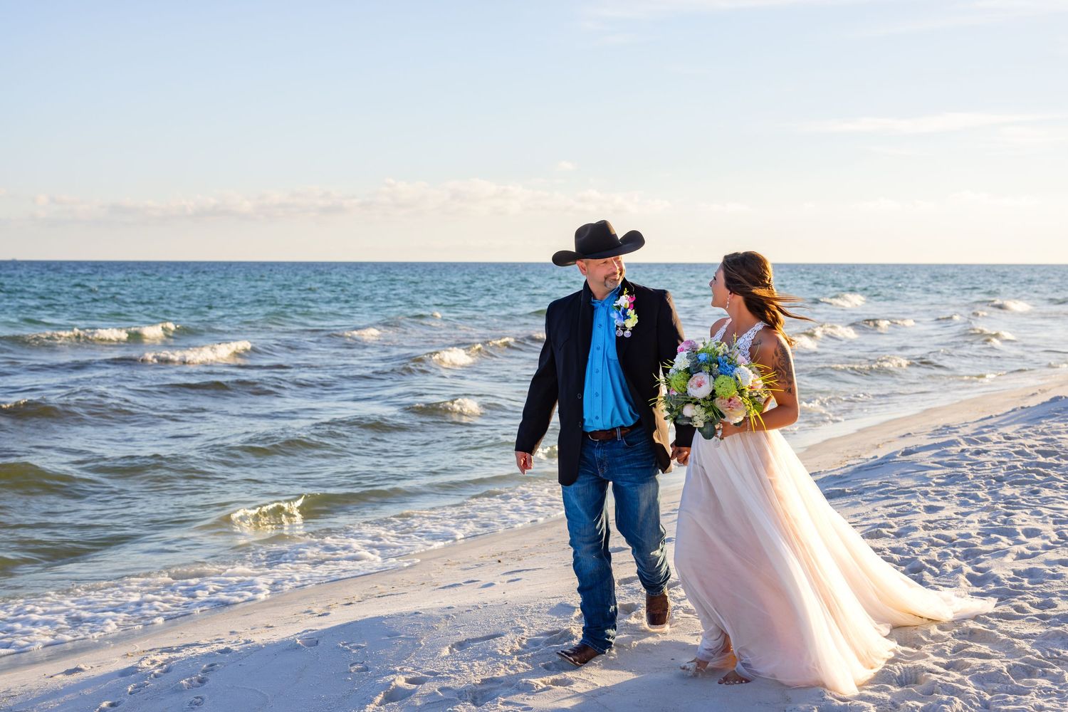 Newlyweds walk along shoreline during beach wedding with waves crashing behind them.