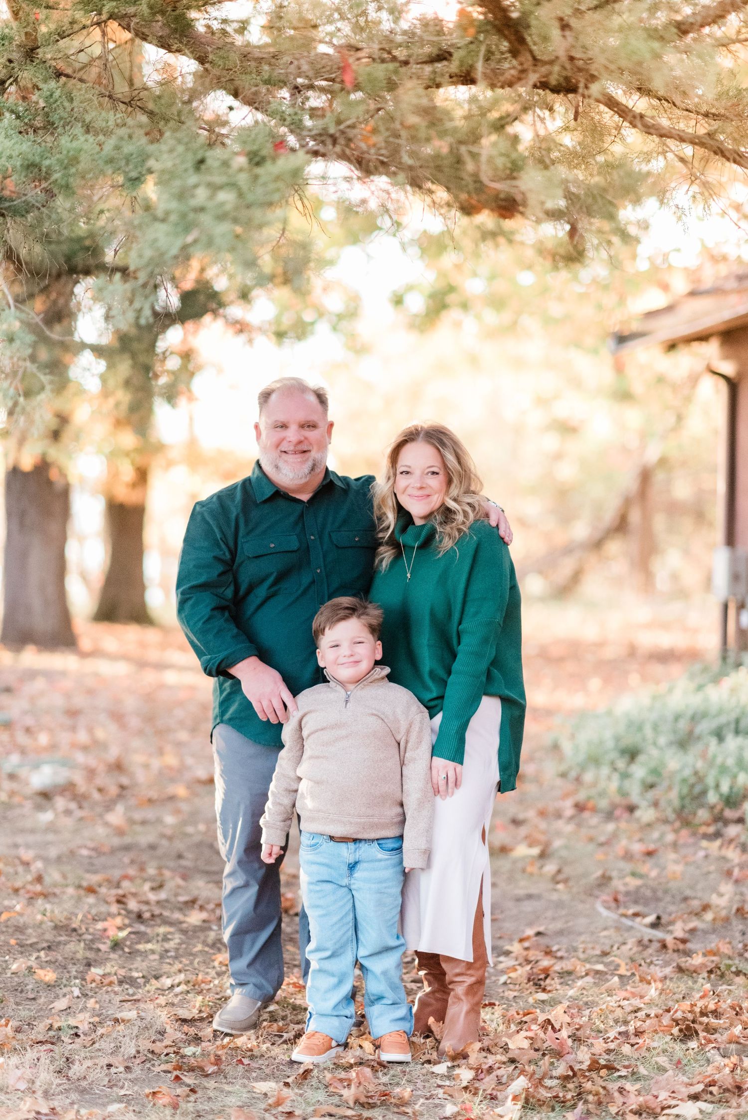 A small family poses together outdoors during fall wearing coordinated green and neutral outfits.