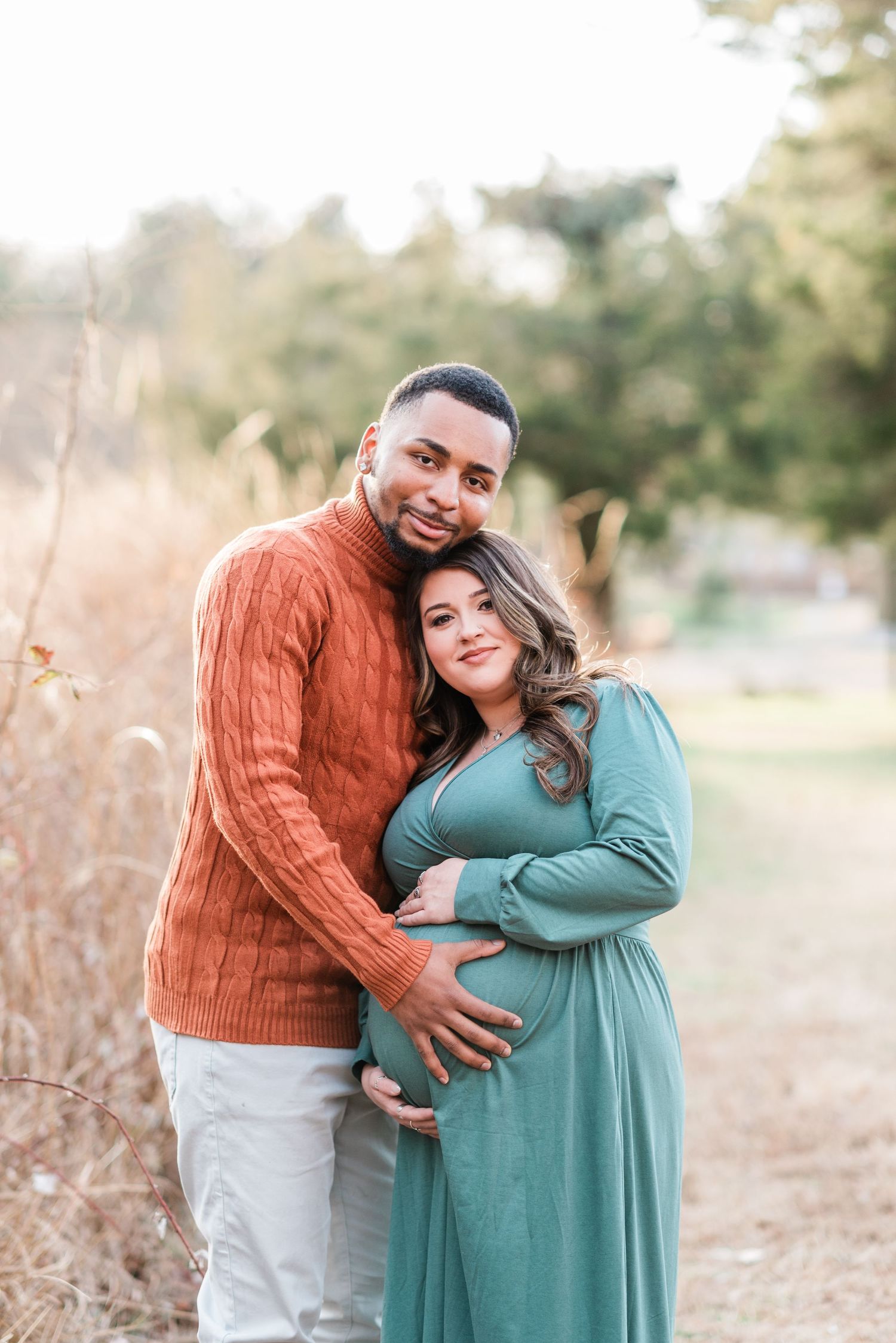 Maternity photo of couple embracing in fall field, wearing teal dress and orange sweater.