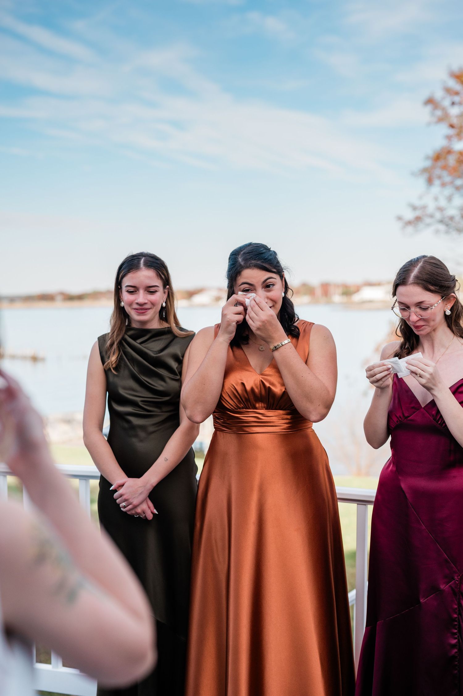 A group of bridesmaids in burgundy and brown dresses share a candid laughing moment together on a sunny day.