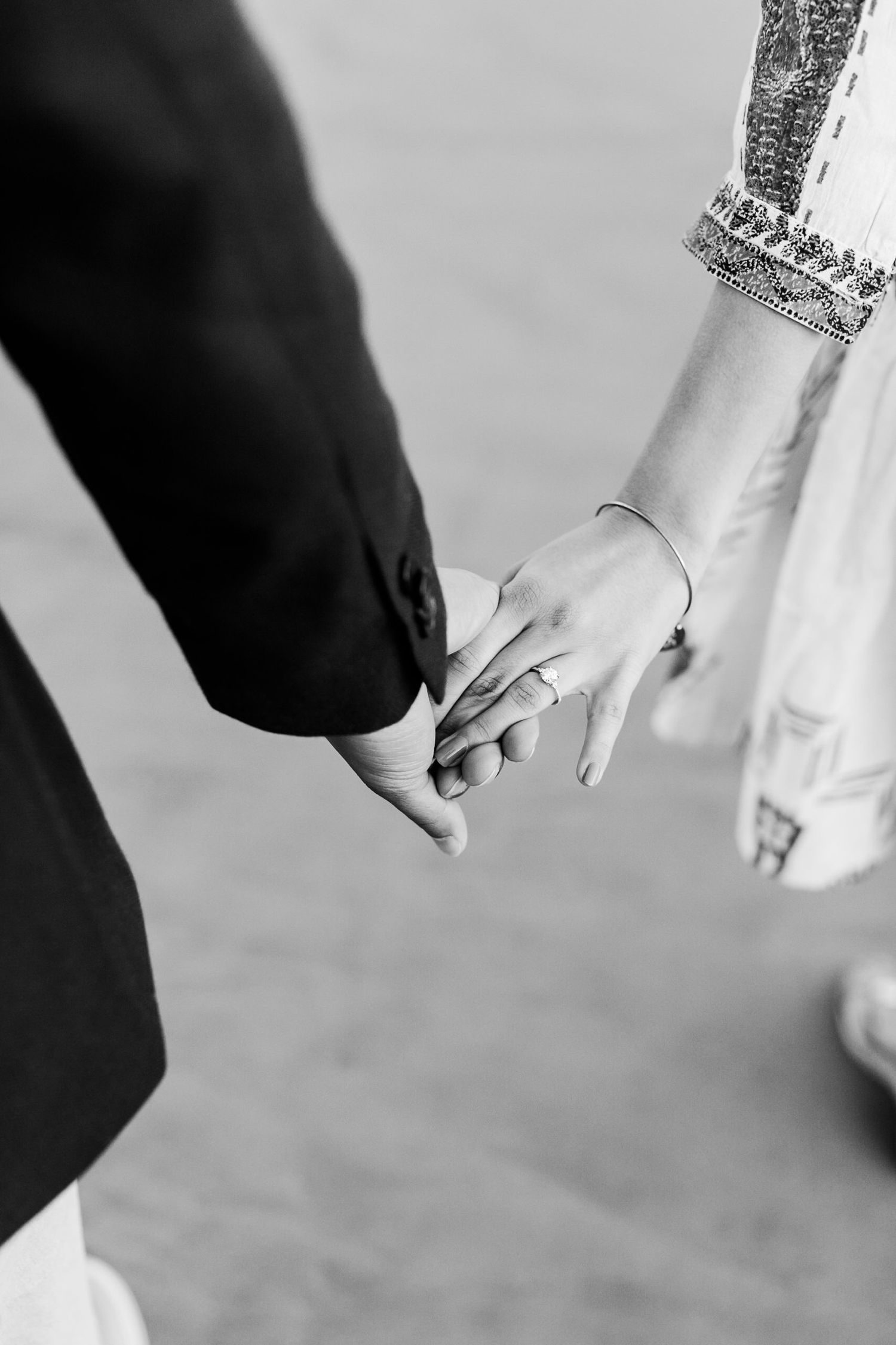 Black and white photo of bride and groom holding hands, woman wearing wedding ring and embroidered dress.