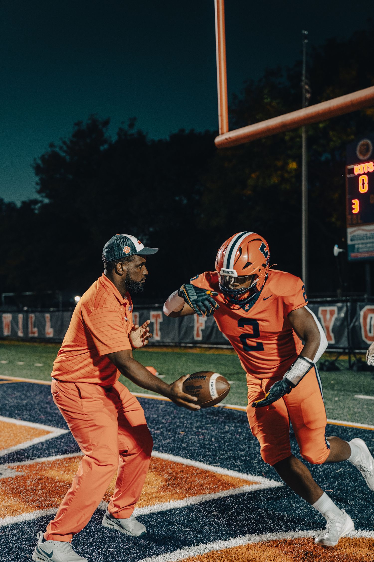 Football player and coach interact on field at night in orange uniforms.