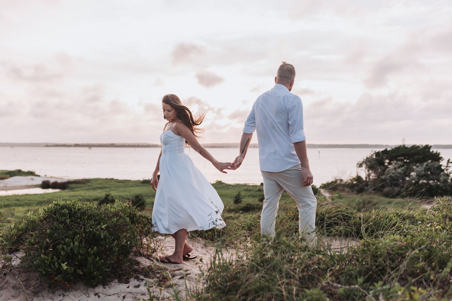 A couple walks hand in hand along a coastal path at sunset surrounded by marsh grasses.