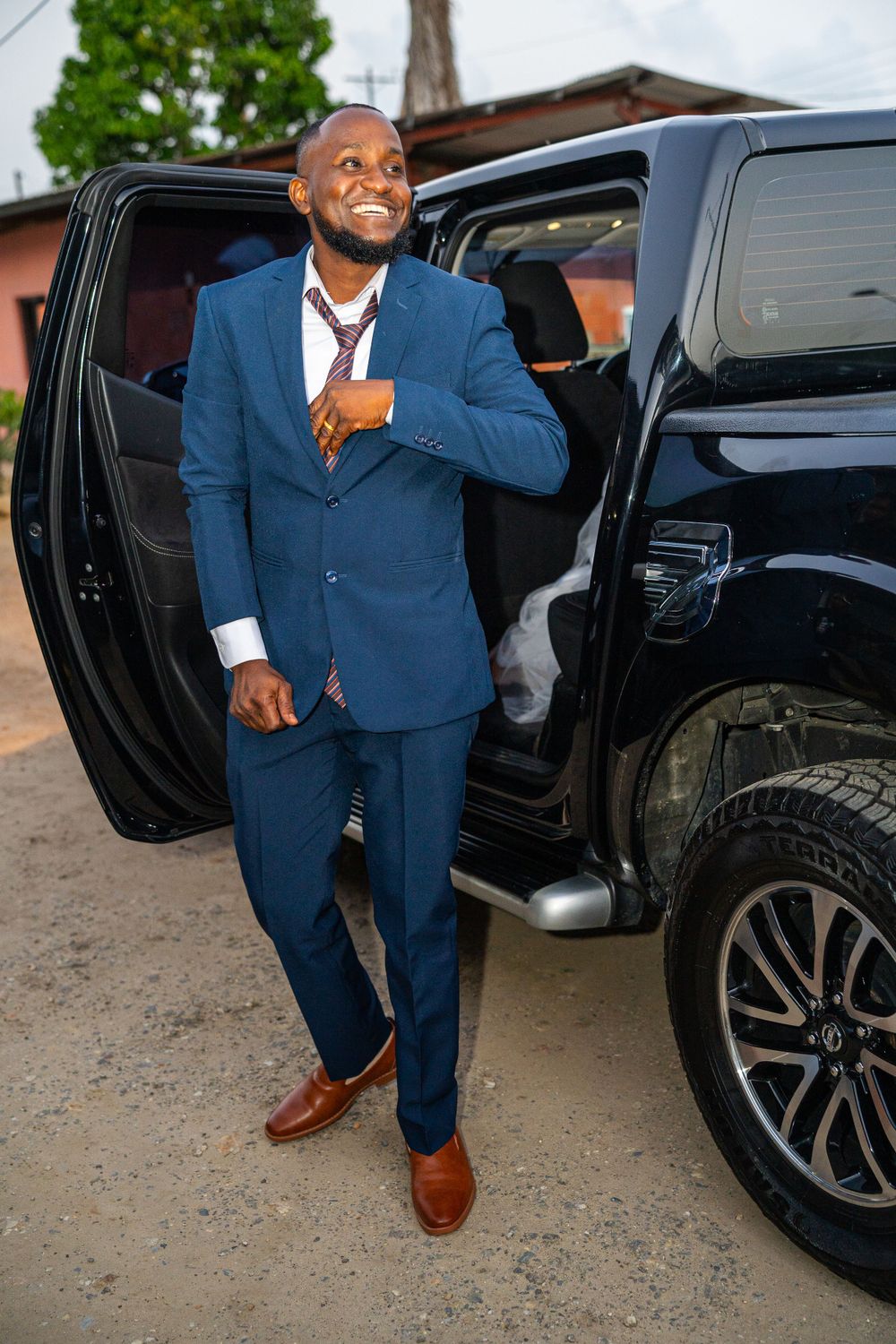 Groom in navy blue suit standing by black SUV with door open.