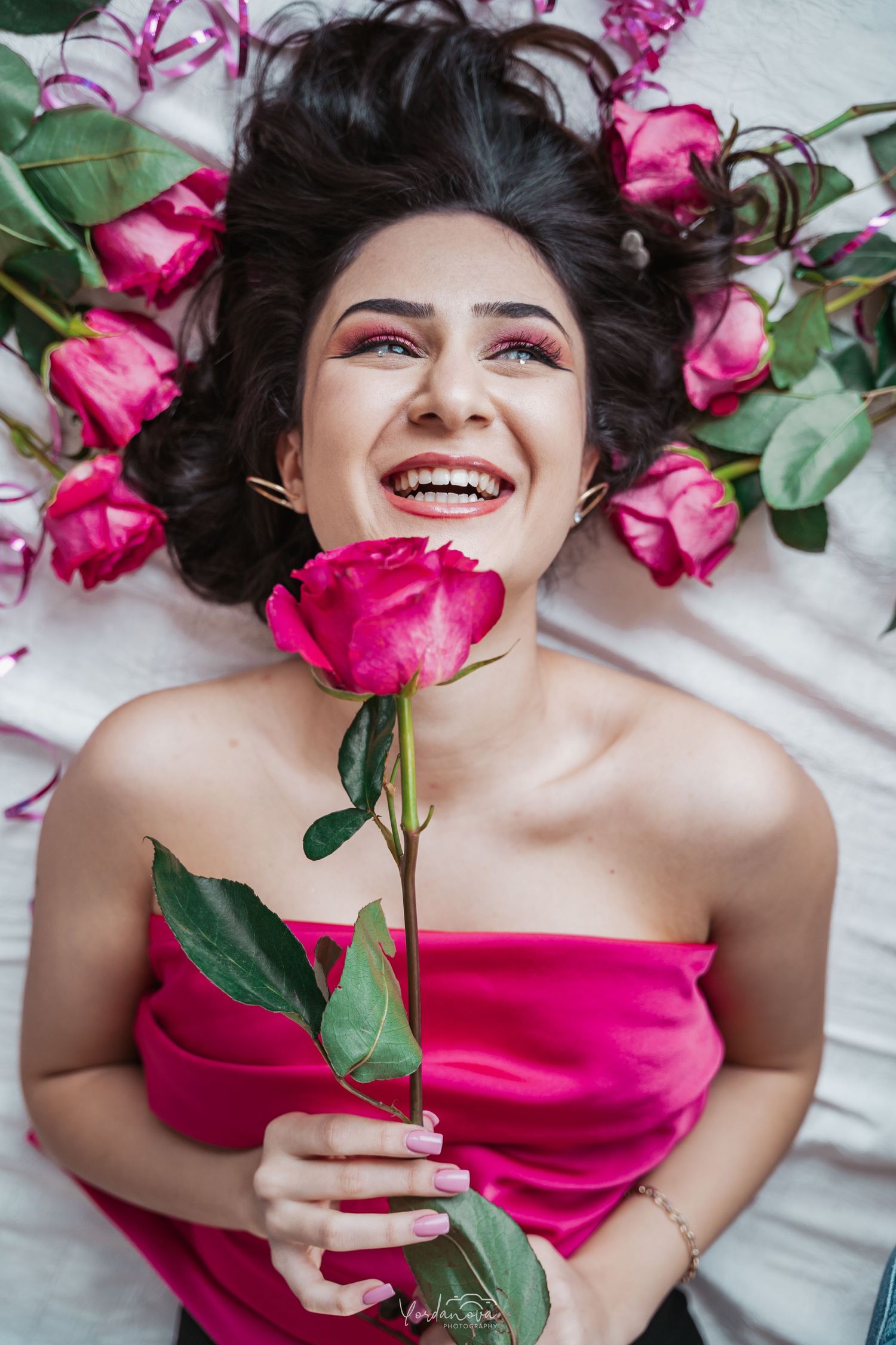 Woman in pink dress lying among vibrant pink roses and greenery smiles joyfully while holding a single rose.