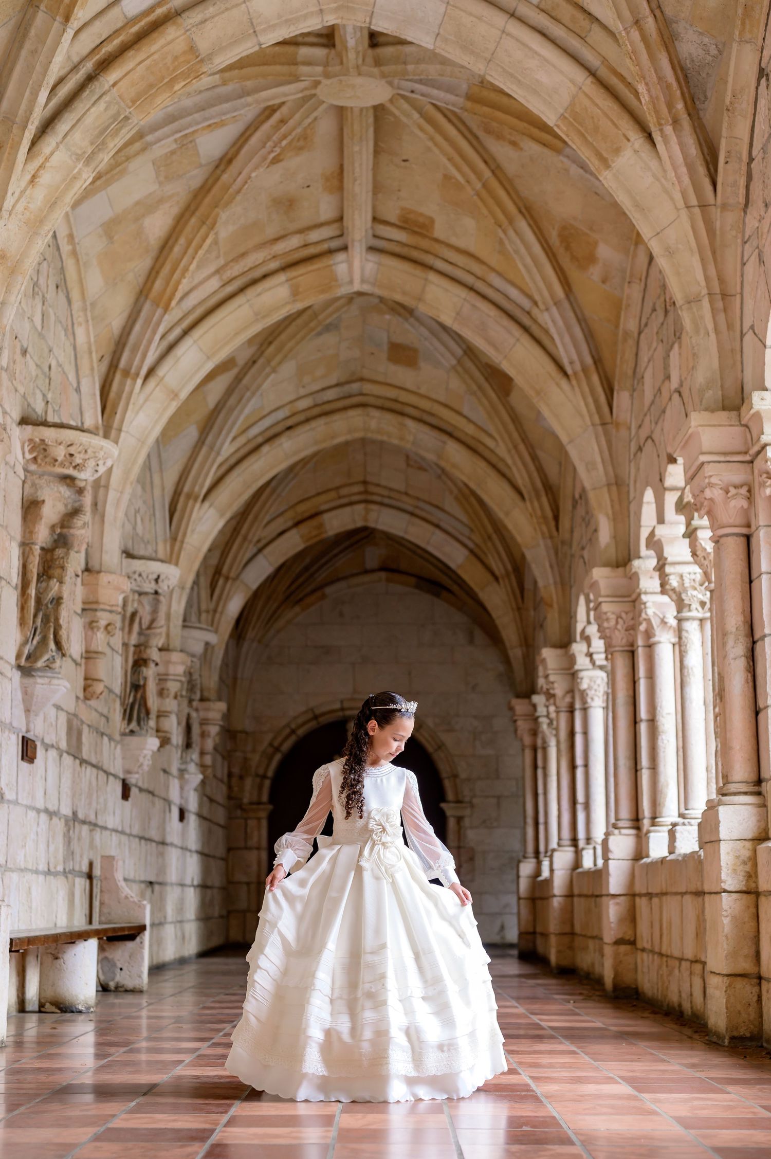 A girl in an elegant white communion ball gown poses in a stunning gothic stone archway corridor with vaulted ceilings.