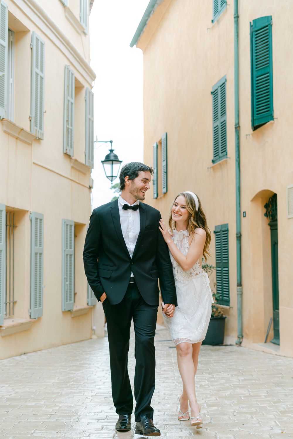 A couple in formal attire walks hand in hand down a charming Mediterranean street with turquoise shutters and cream walls.