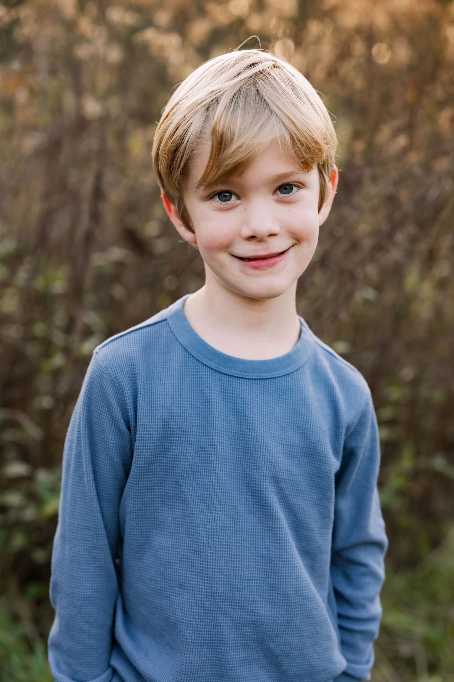 Young child wearing a blue thermal long sleeve shirt smiles warmly while standing outdoors against a natural background.