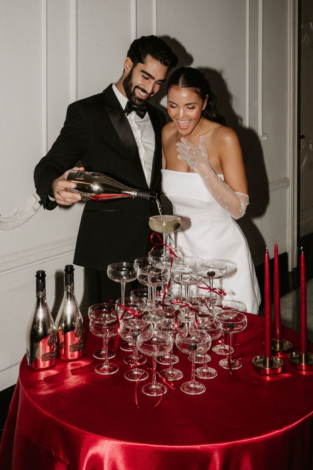 Couple pouring champagne into pyramid of glasses on red tablecloth at wedding celebration.
