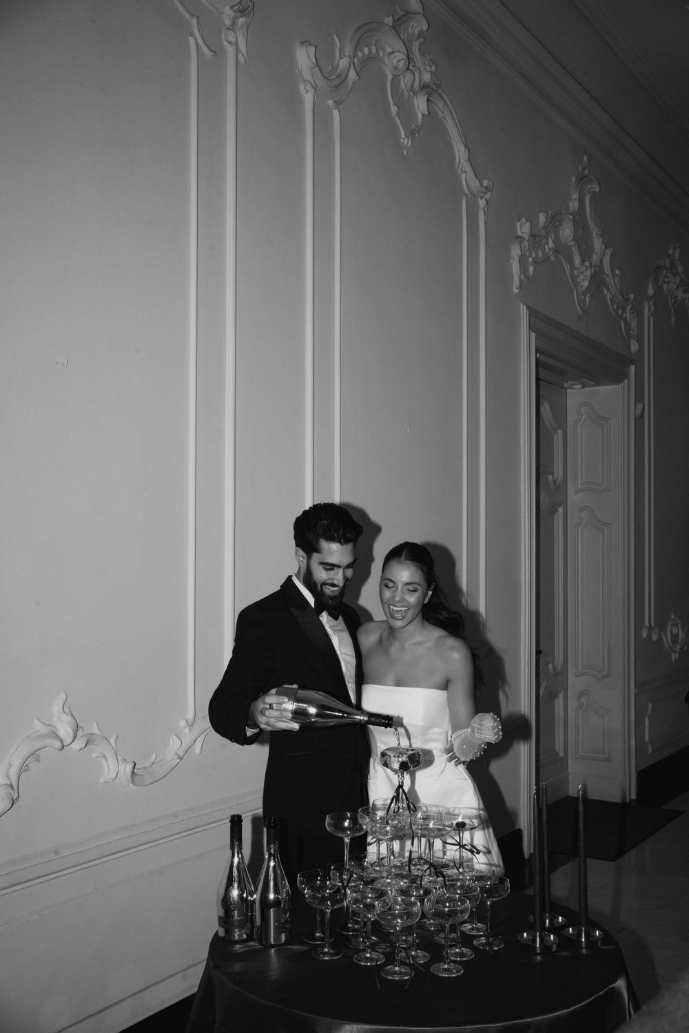 Black and white photo of couple pouring champagne into tower of glasses.