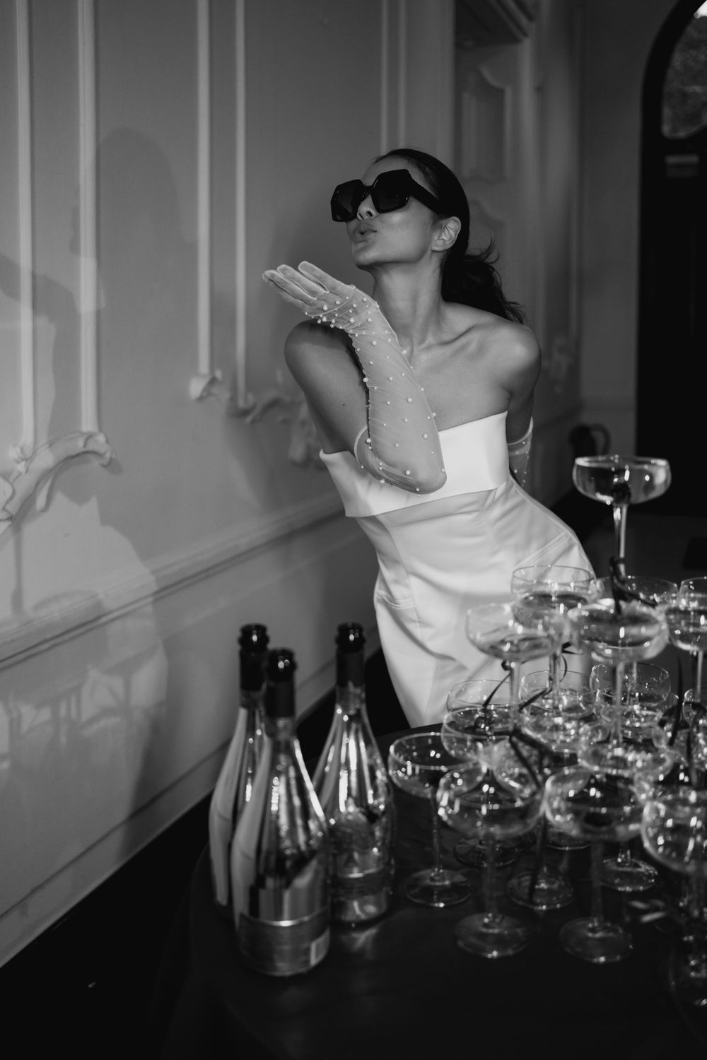 Bride in sunglasses posing playfully by champagne tower in elegant room.
