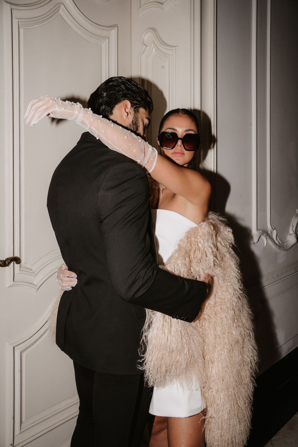 An elegant couple embraces while wearing formal attire and a feathered coat in a classic interior setting.