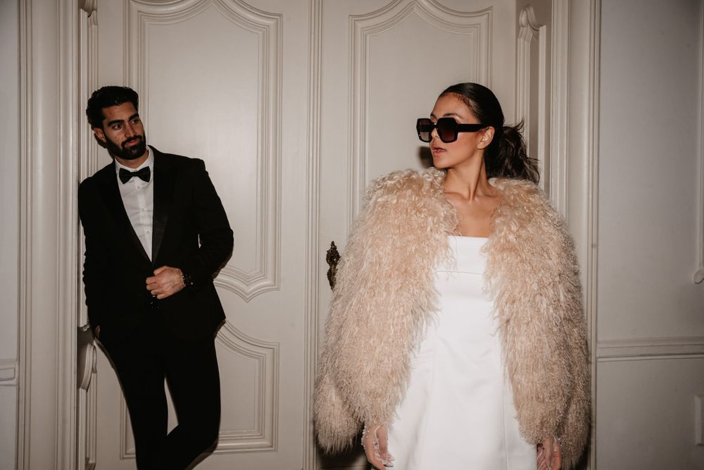 A stylish couple poses in formal attire with a fluffy beige coat and classic tuxedo against an ornate white wall.