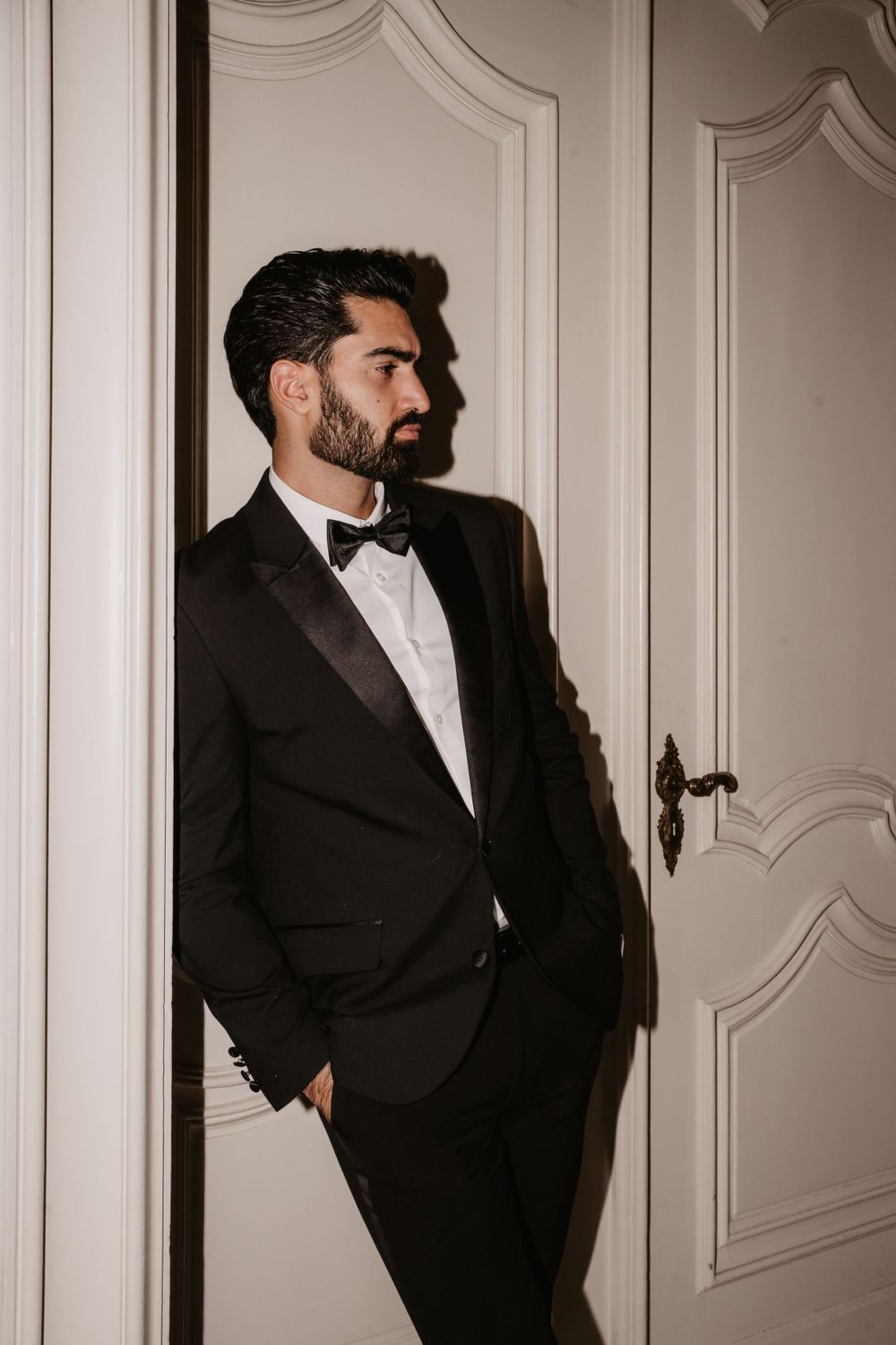 A dapper gentleman in a black tuxedo and bowtie poses against an ornate white wall.