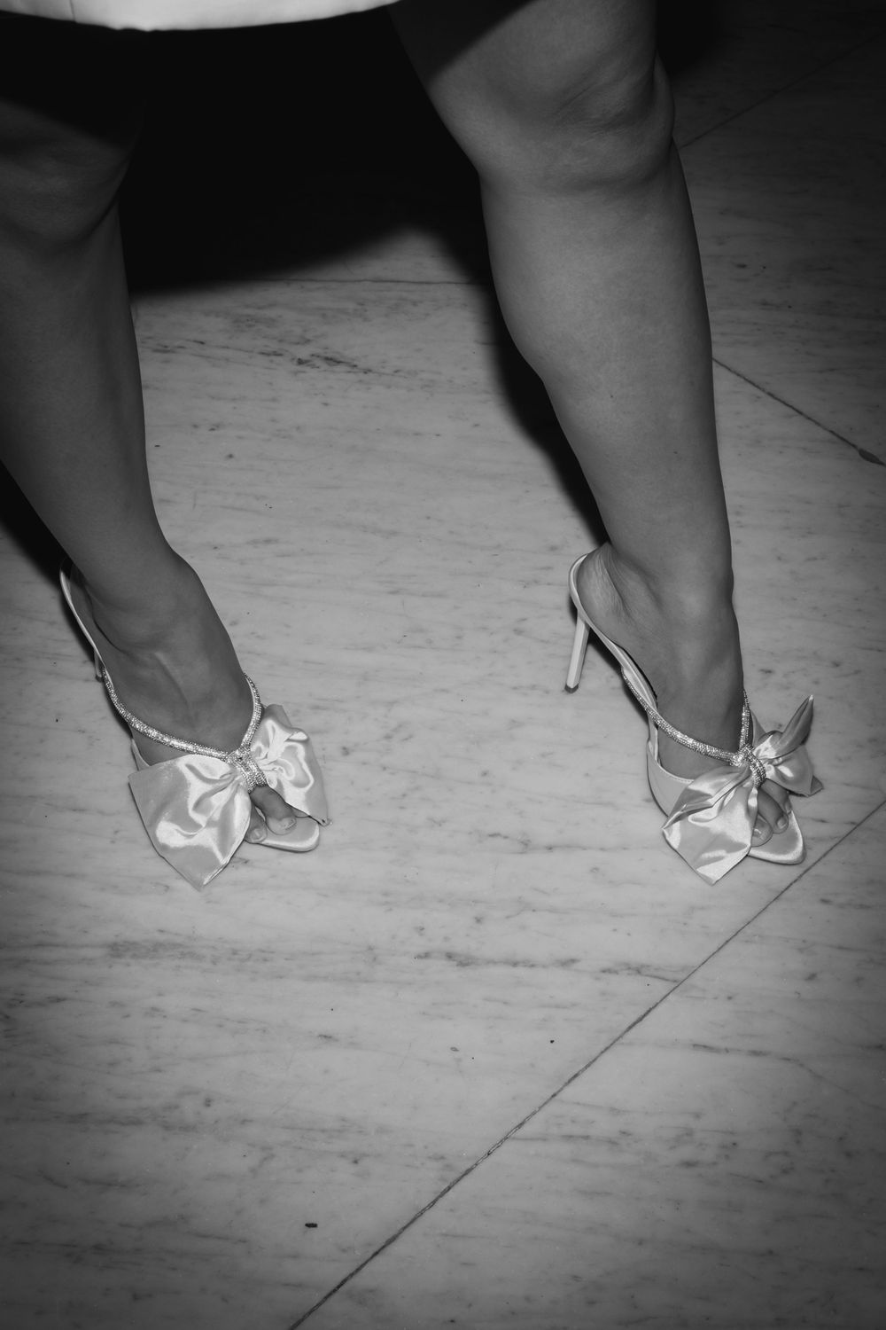 A pair of white satin bow heels on marble flooring captured in black and white.