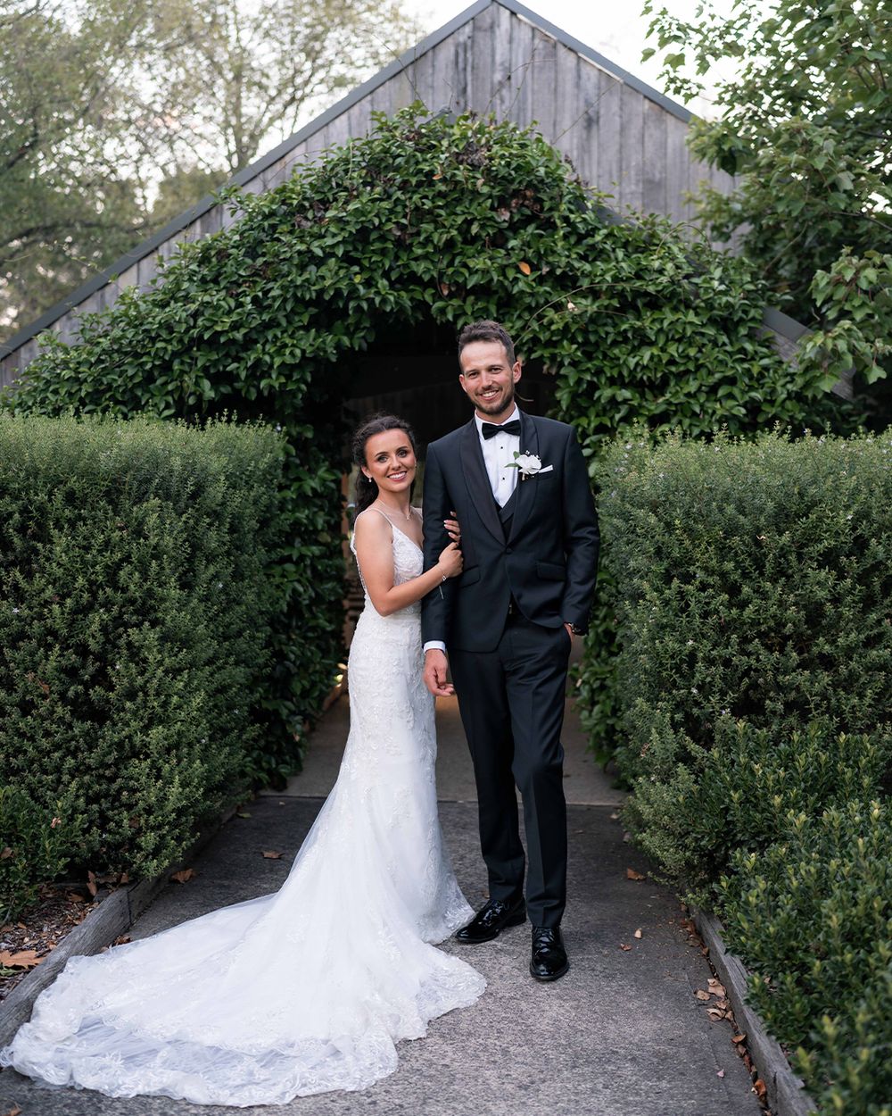 A bride in a white lace gown and groom in a black tuxedo pose together in a lush garden archway on their wedding day.