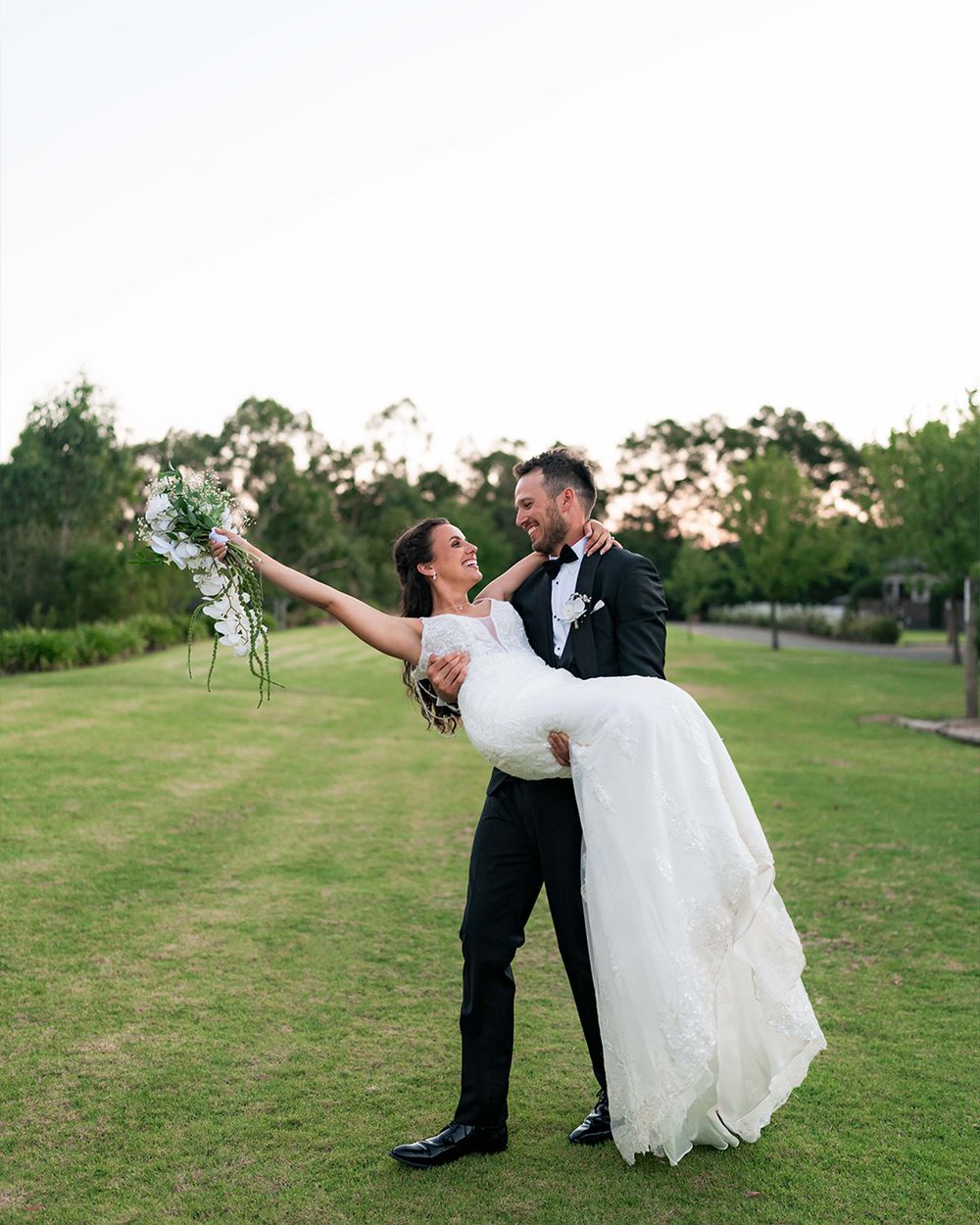 Groom lifts bride in white gown as she raises bouquet on green lawn at outdoor wedding celebration.