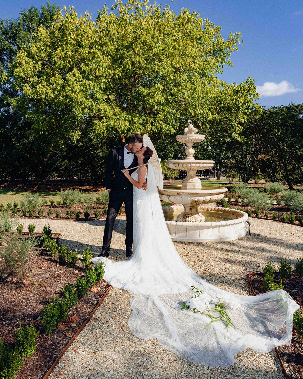 Bride and groom kissing near a stone fountain in a lush garden, with a long flowing white wedding veil trailing on the ground.