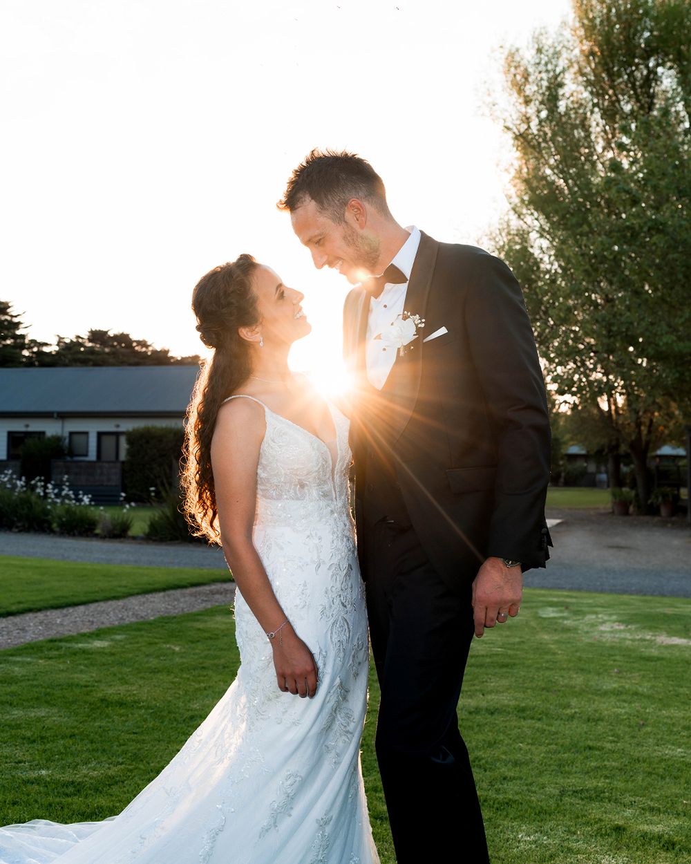 Bride and groom share a romantic sunset moment outdoors, golden light glowing between them on lush green lawn.