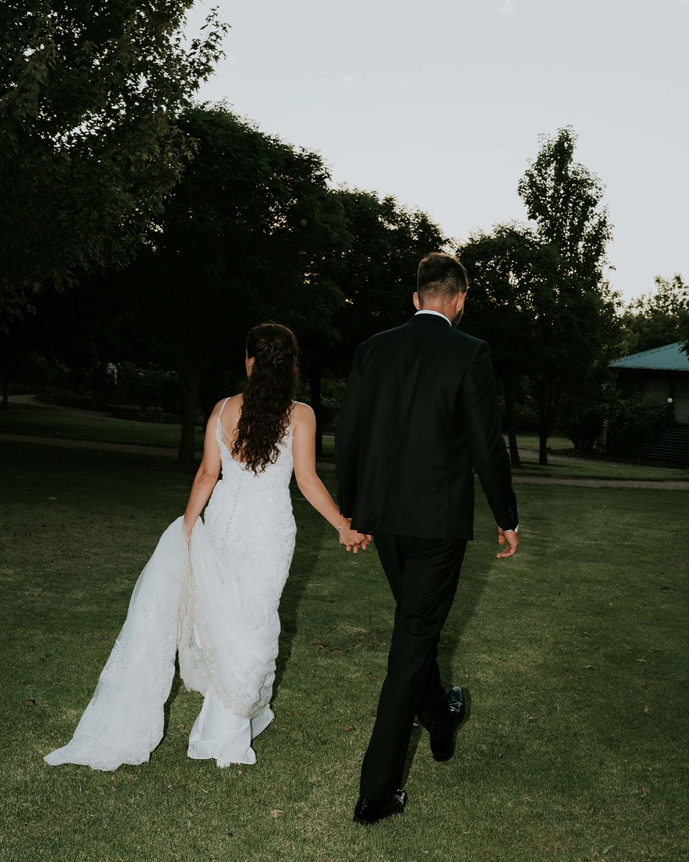 Newlywed couple walking hand in hand across green lawn at dusk, bride in white gown and groom in black suit.