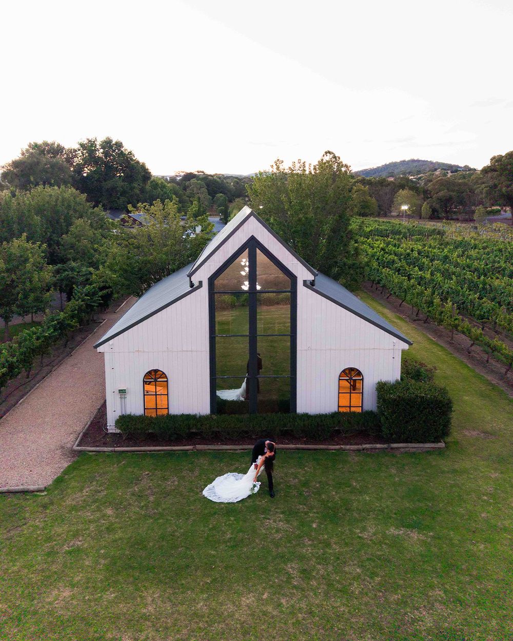 Aerial view of couple kissing outside a white chapel wedding venue surrounded by lush vineyards at dusk.