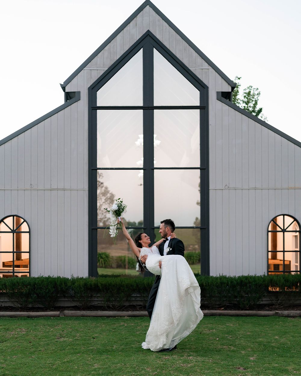 Groom lifting bride in front of modern white barn wedding chapel with large glass windows at dusk.