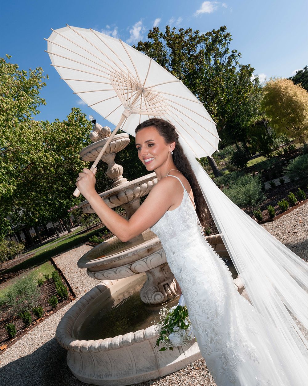 Bride in white gown holding parasol umbrella beside stone fountain in garden on sunny wedding day.