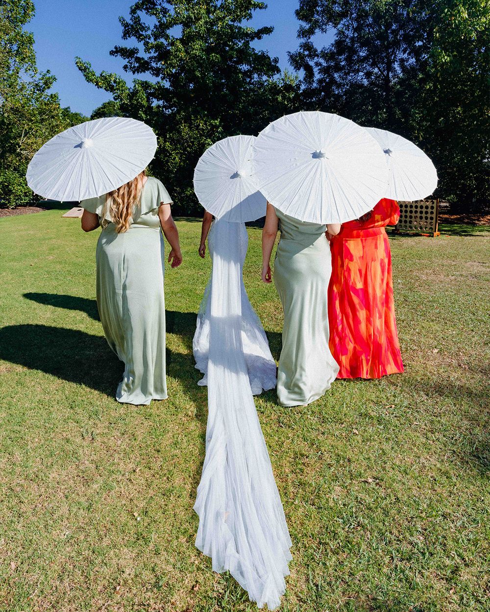Bride and bridesmaids holding white parasols outdoors, wearing white and sage green gowns on green grass.