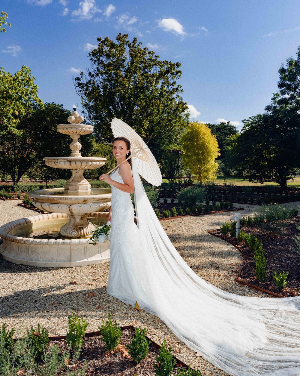 Bride in white gown holding parasol stands by stone fountain in lush garden on sunny day.