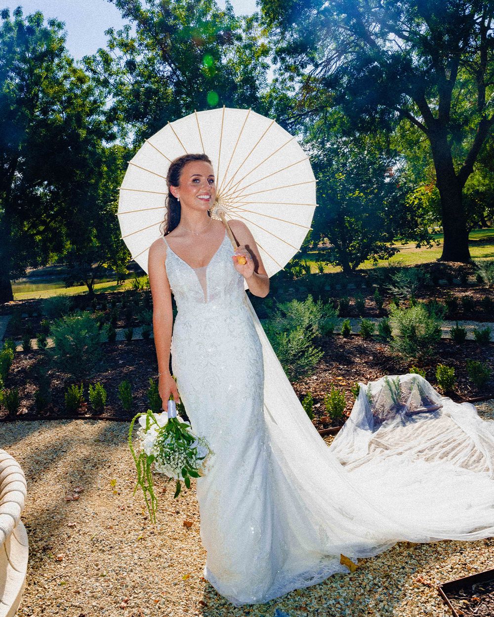 Bride in white lace gown holding parasol and bouquet in sunny garden setting.