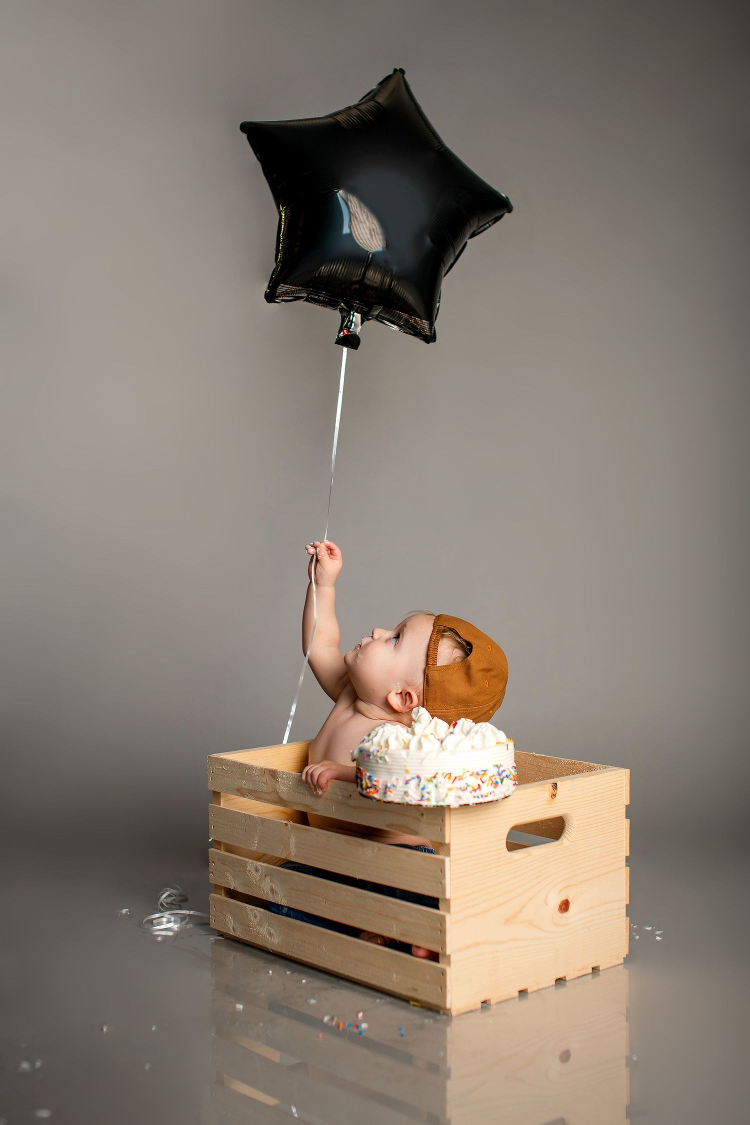A studio photo showing a baby sitting in a wooden crate during a creative cake smash photoshoot with balloons and bubbles.