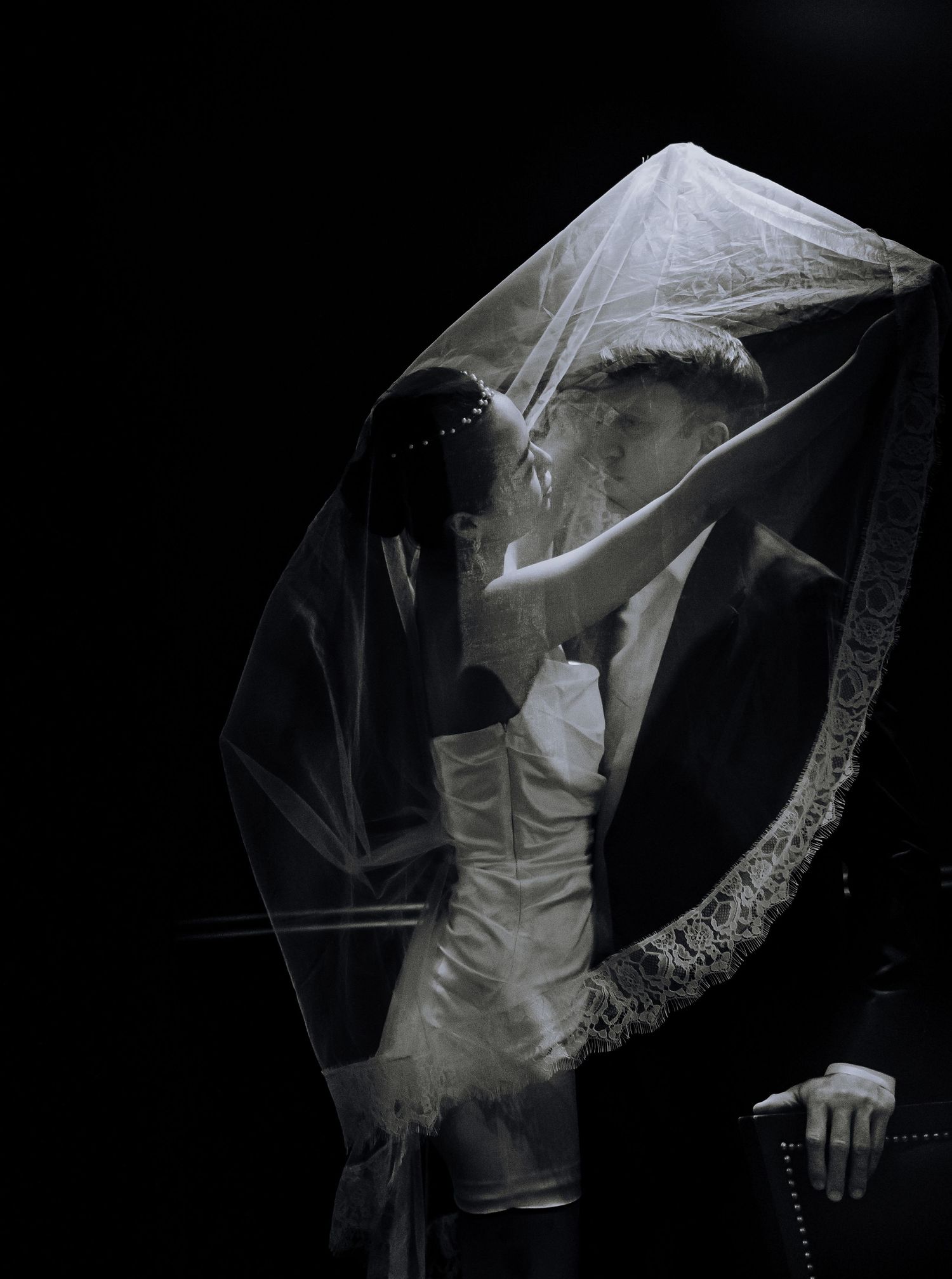 Romantic black and white artistic photo of a couple embracing under a sheer veil against dark background during wedding ceremony.