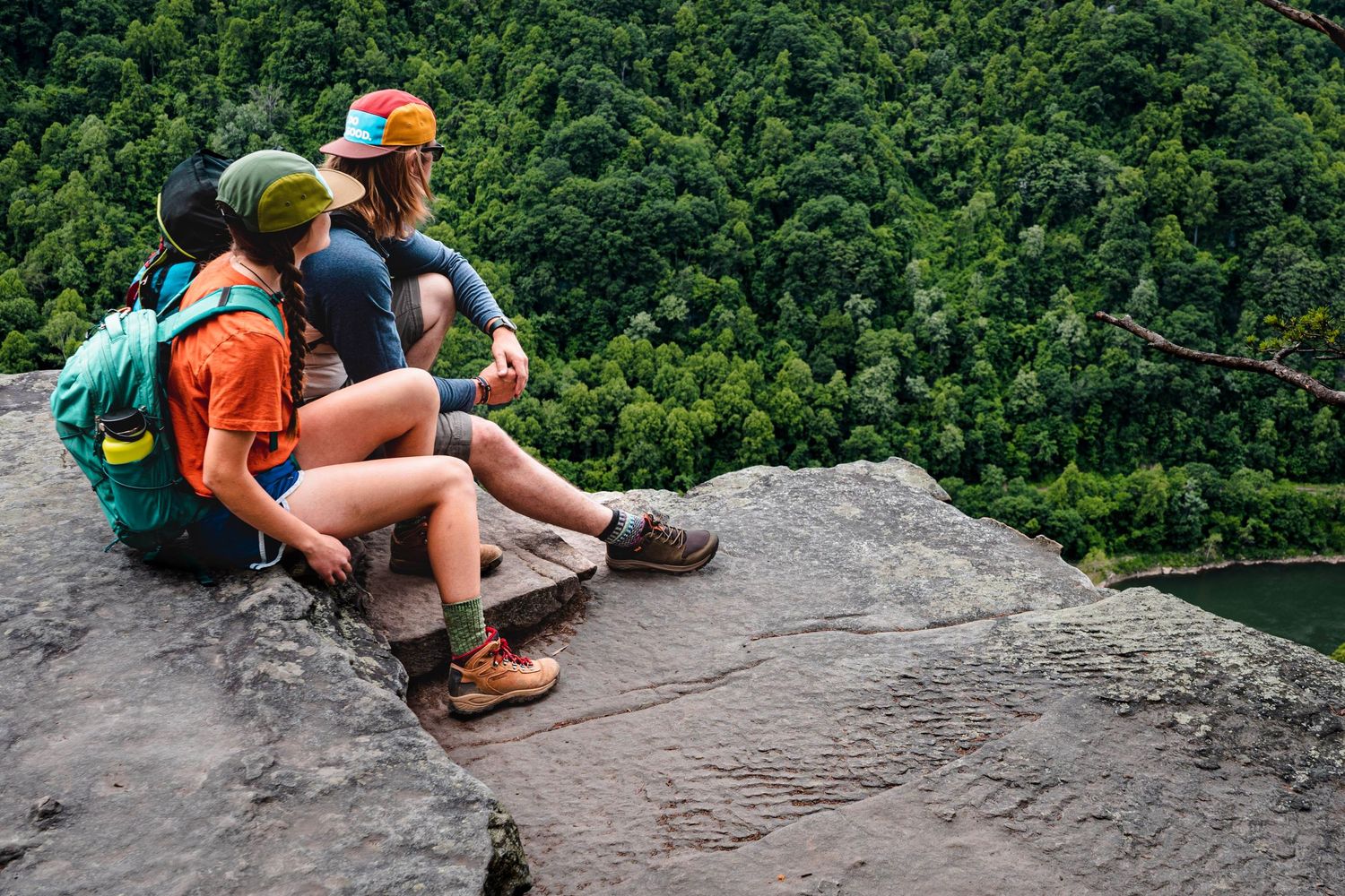 a couple sitting atop a mountain, starting out over a river and into the vast green trees at new River gorge national park in west virginia