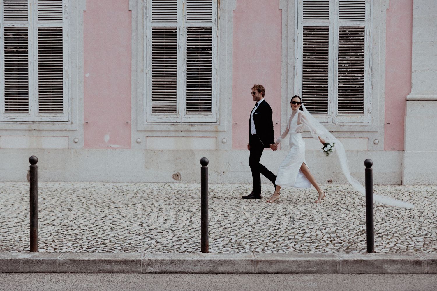 A bride and groom walk together along a cobblestone sidewalk next to a classic pink building with white shuttered windows.