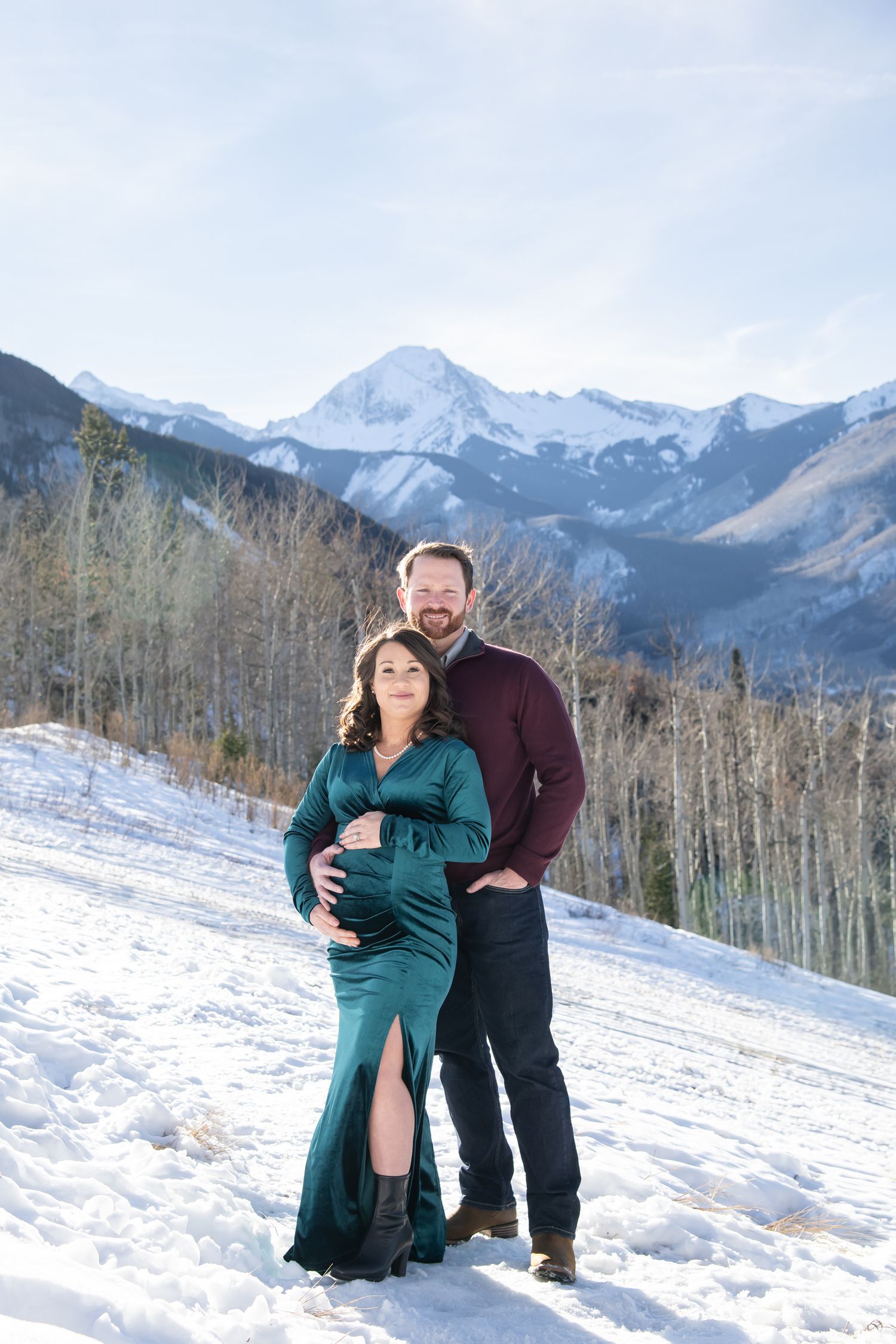 A couple poses together in the snow with dramatic mountain peaks and aspen trees in the background for maternity photos in Aspen Colorado
