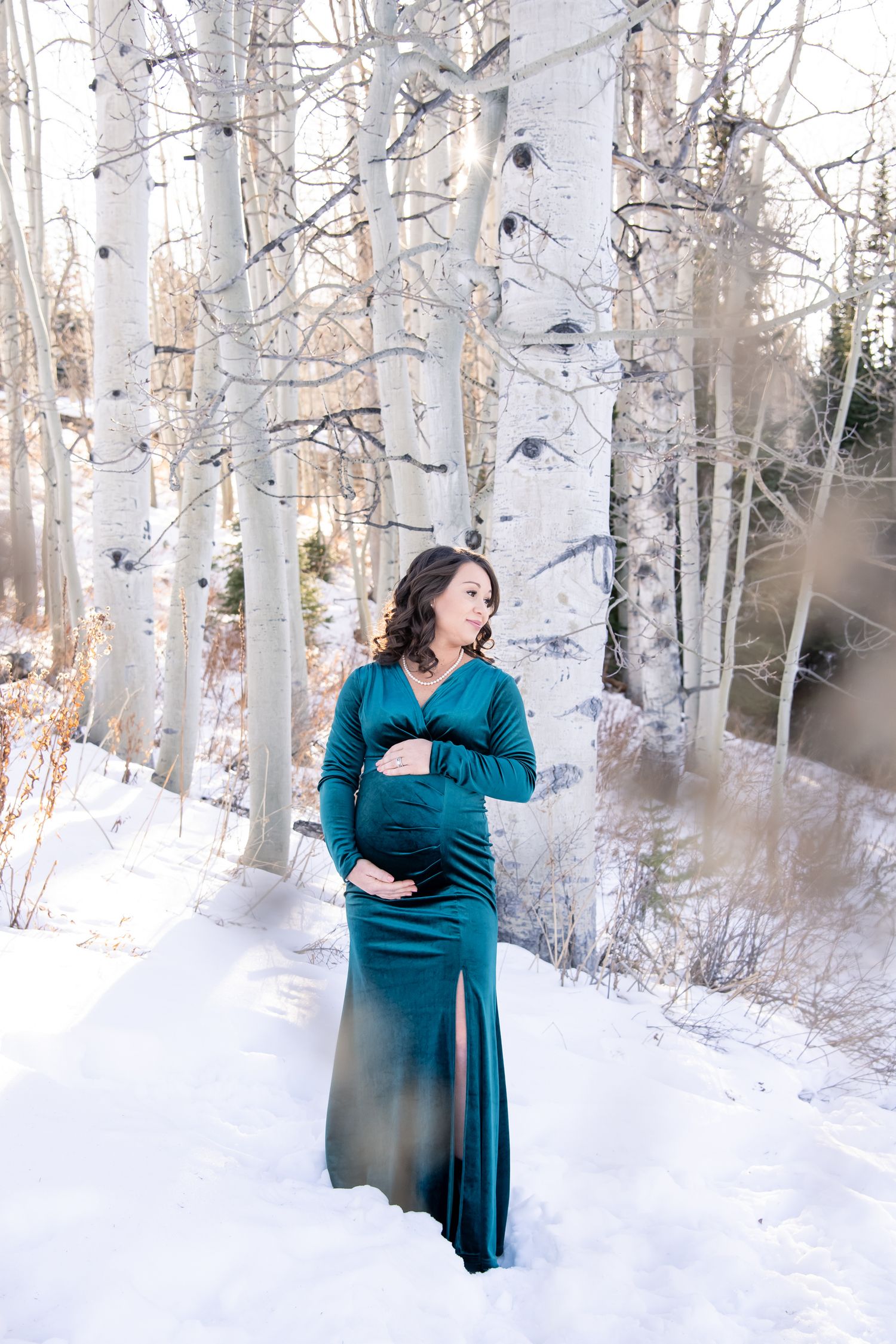 A beautiful woman stands in the snow with aspen trees behind her during a maternity photo session in Aspen Colorado