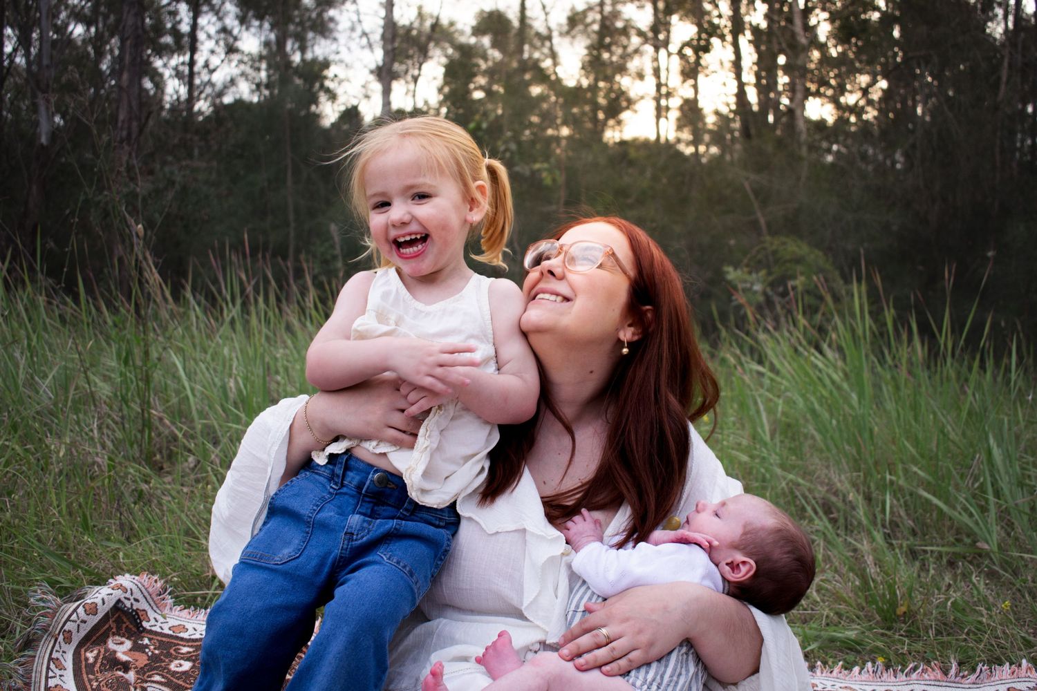 mum tickling toddler as she holds a newborn, both laughing, in a grassy field, during a newborn photoshoot Maitland NSW