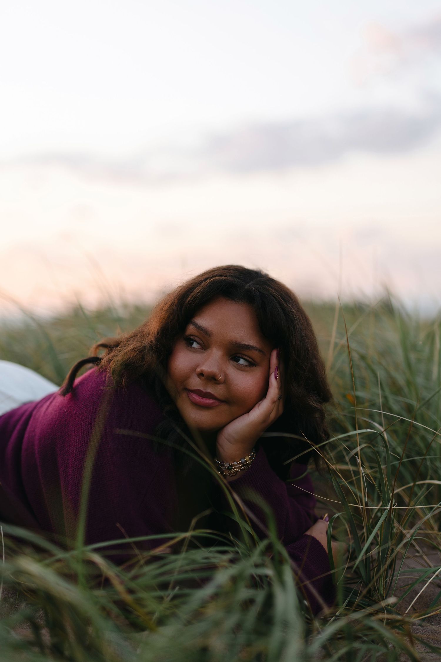 A person in a burgundy sweater lies in tall grass during sunset with a dreamy expression.